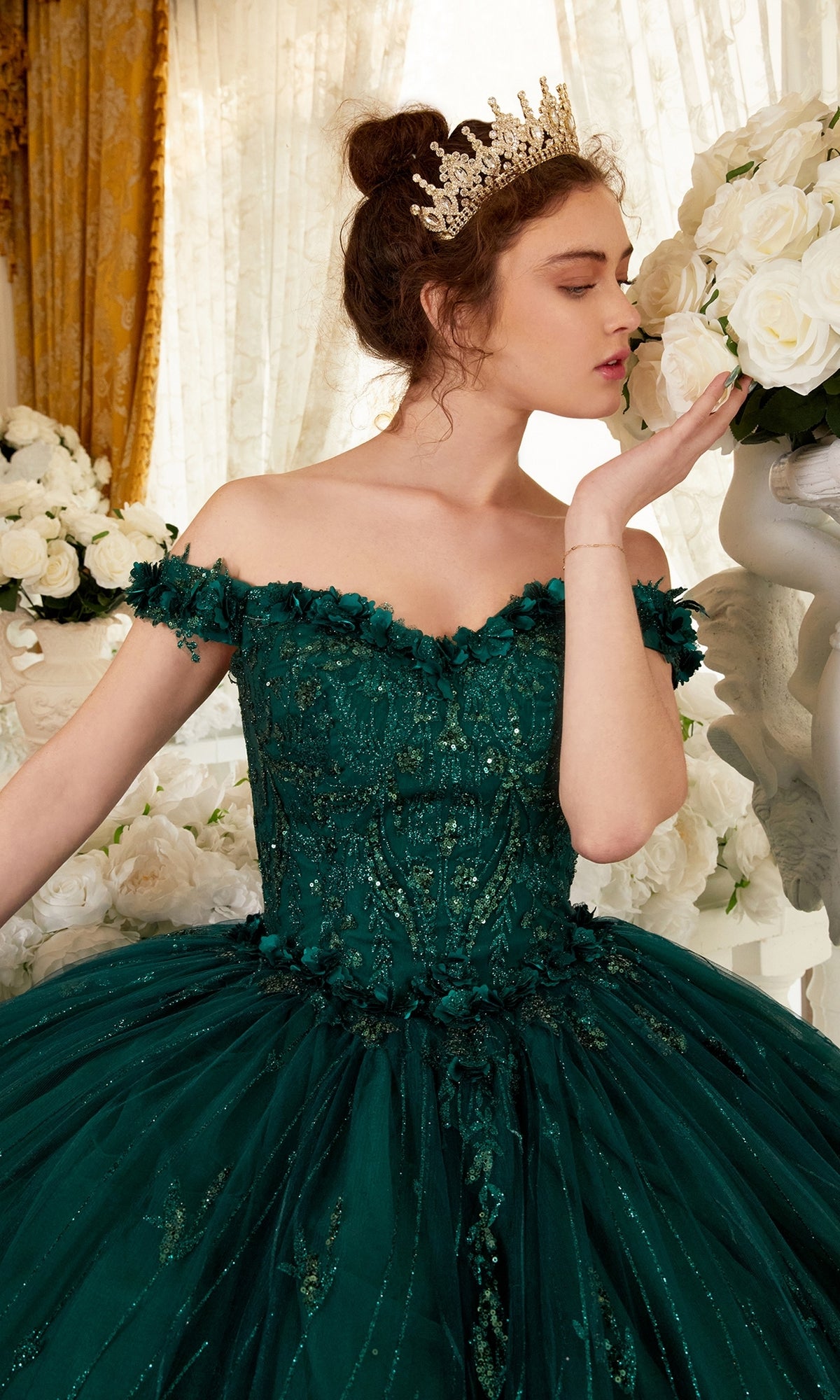 A woman in the Ladivine 15704 Off-the-Shoulder Quince Ball Gown and a tiara stands among white roses, holding one flower close to her face.